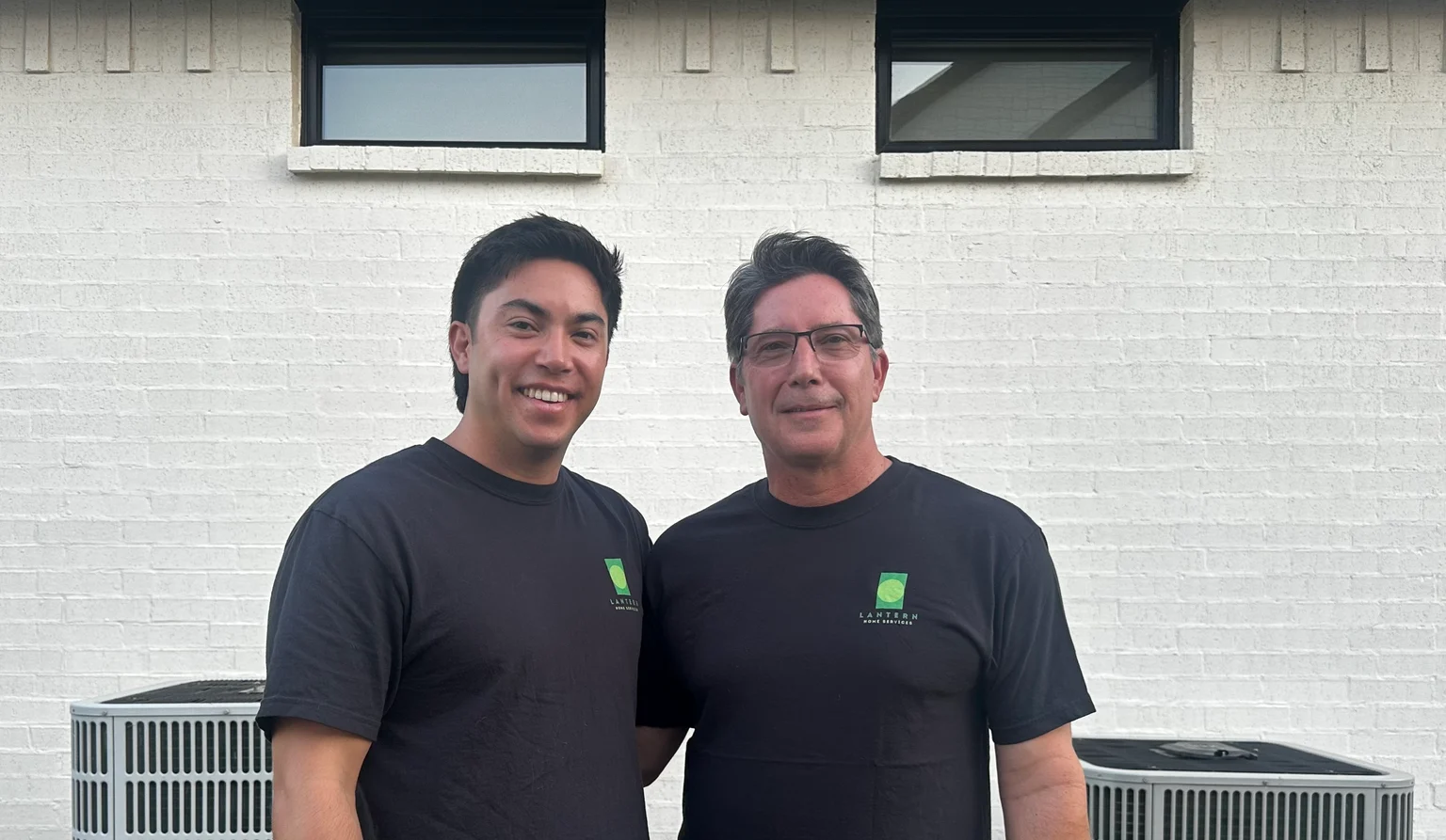 Jay and Daryl of Lantern Home Services in branded shirts, standing between two air conditioning condensers outside a Dallas-Fort Worth home